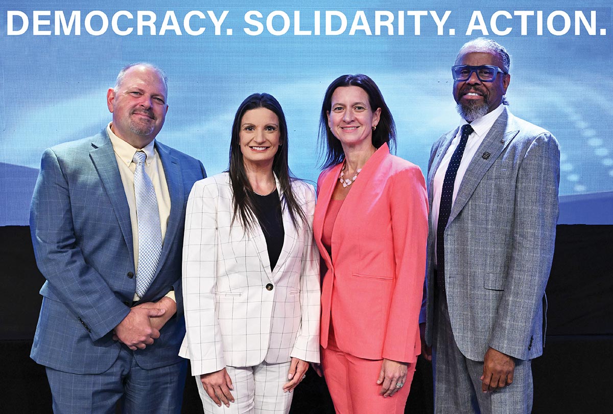 Four people in business attire pose in front of a blue backdrop with the text 