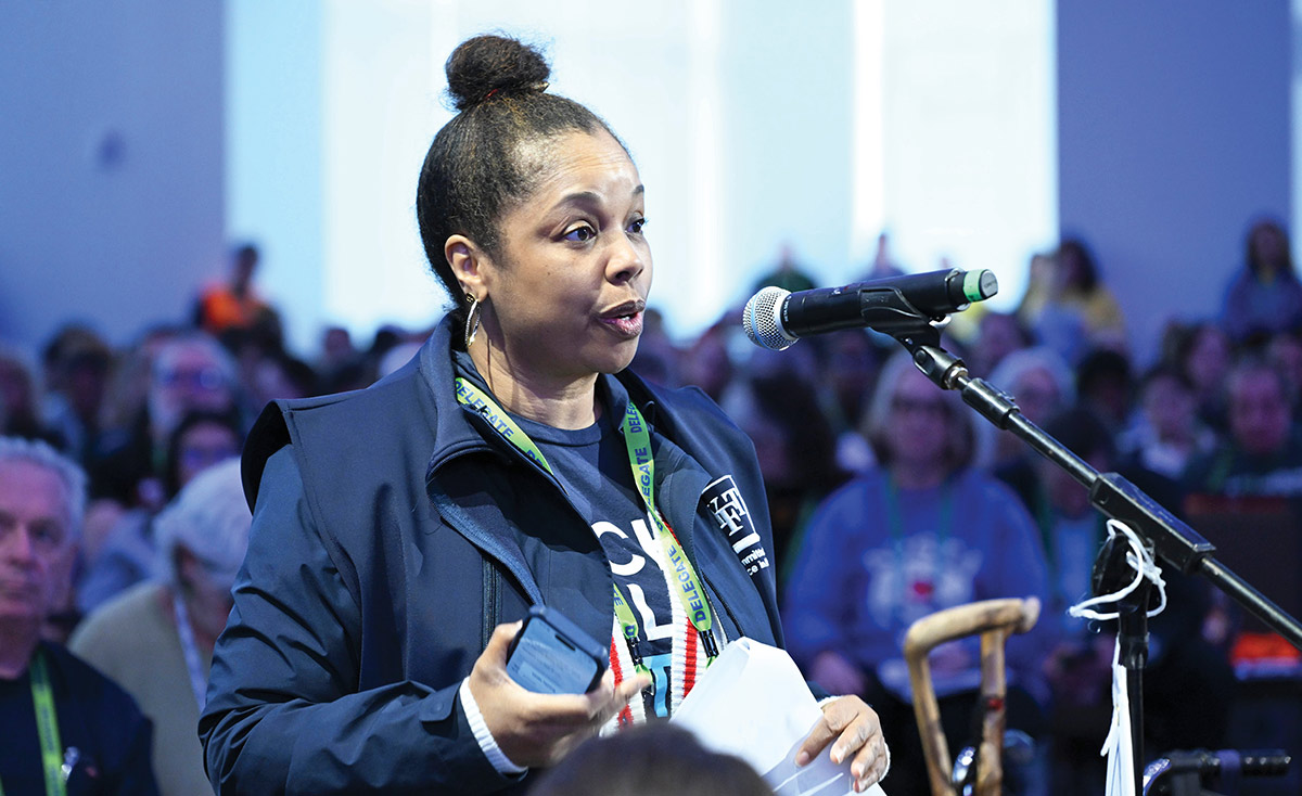 A woman wearing a "DELEGATE" lanyard speaks into a microphone at a large event. She holds a smartphone and papers while addressing the assembly, with a blurred audience in the background.