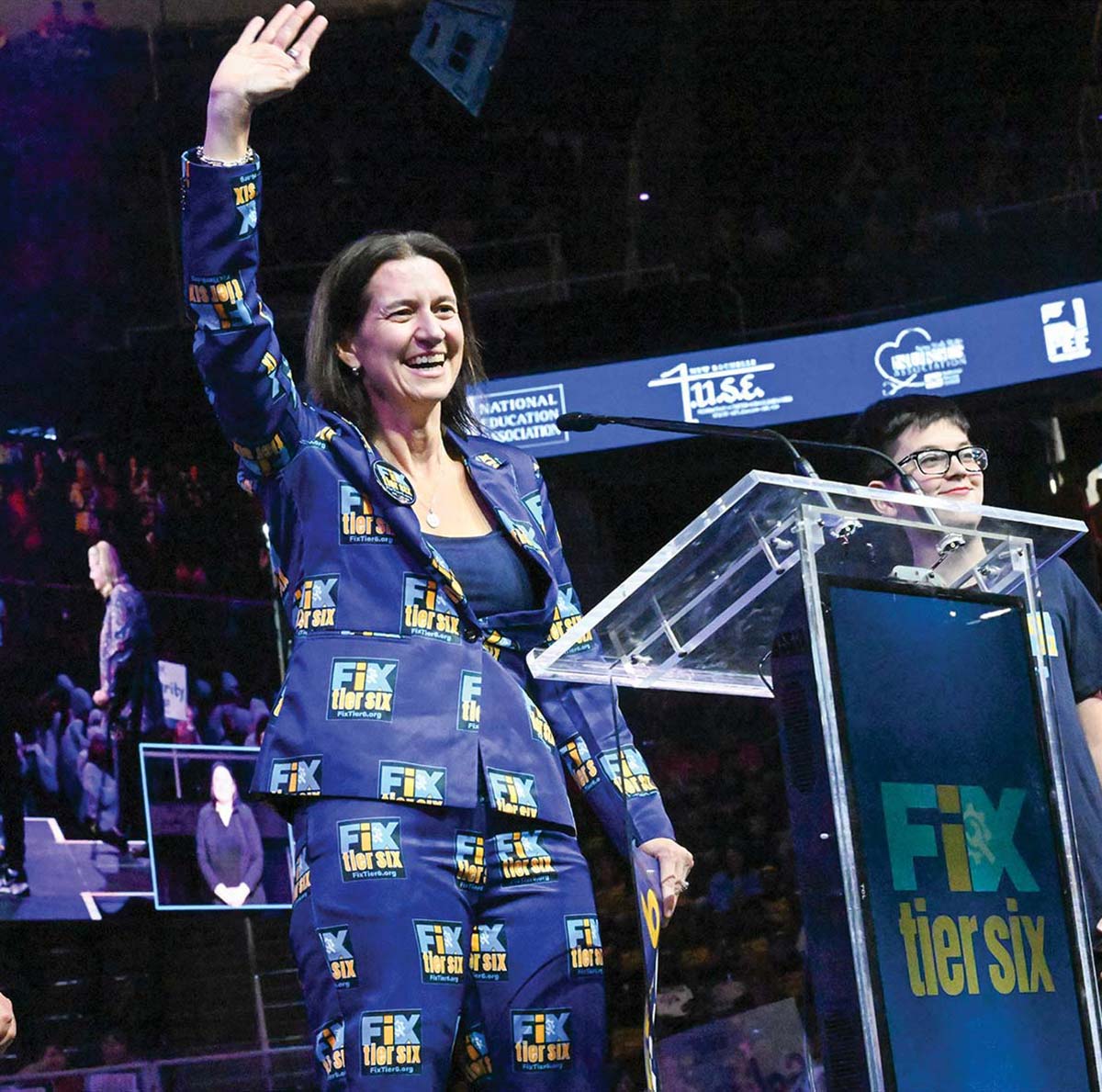 A woman in a blue suit covered in "Fix Tier Six" logos smiles and waves while standing at a clear podium on a stage. The background shows a large arena with National Education Association banners.