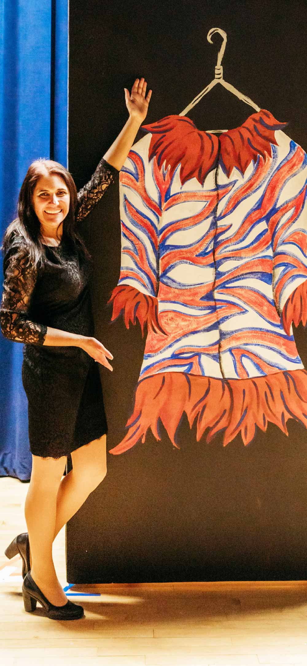 A smiling woman in a black lace dress stands next to a large painting of a colorful, patterned coat on a hanger against a black background.