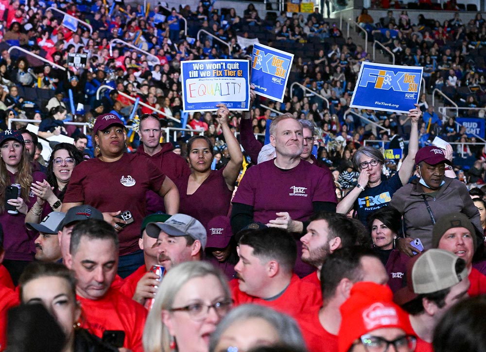A diverse crowd of people in a stadium. Several individuals hold signs that say “We won't quit till we Fix Tier 6! EQUALITY” and “FIX Tier SIX.” Many attendees are wearing maroon shirts.