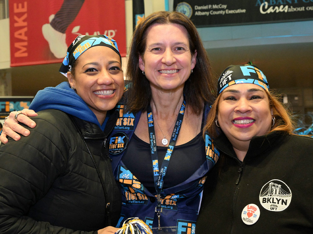 NYSUT President Melinda Person smiles between two women. All three wear “Fix Tier 6” branded apparel, including bandanas and lanyards.