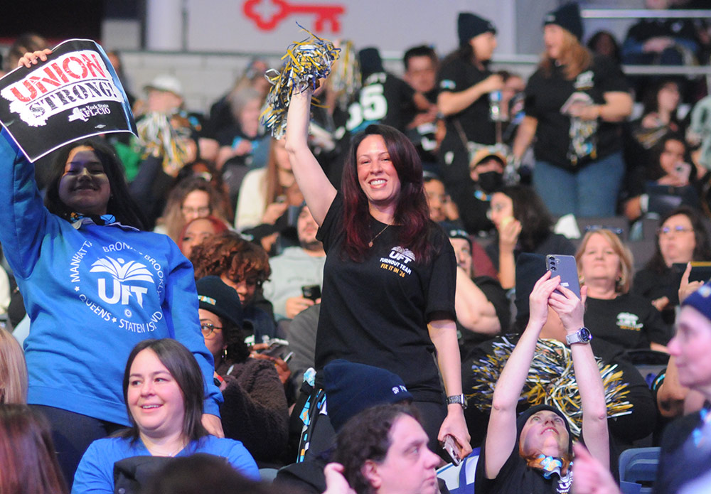 A woman in a black UFT t-shirt smiles and holds a blue and yellow pom-pom in the air. To her left, another person holds a “UNION STRONG” sign. The background is filled with other rally attendees.