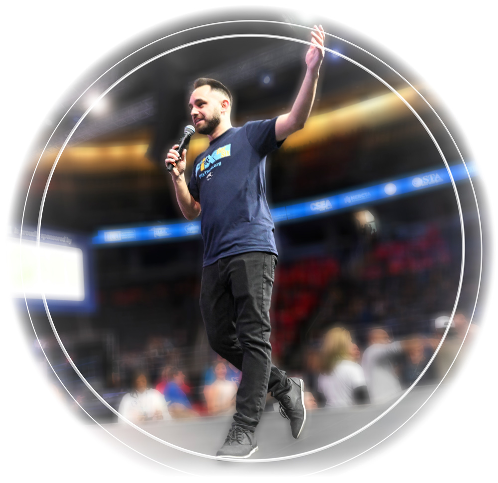 Circular graphic of Gaspare Randazzo on stage, wearing a navy “Fix Tier 6” t-shirt and black jeans. He holds a microphone in one hand and gestures with the other while addressing the stadium crowd.