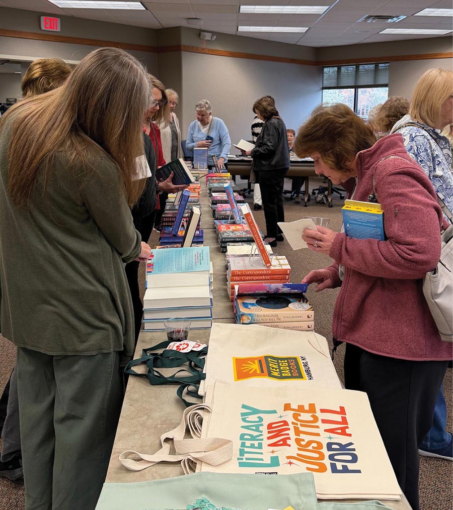 Attendees browsing a long table of books and promotional tote bags at a book fair.