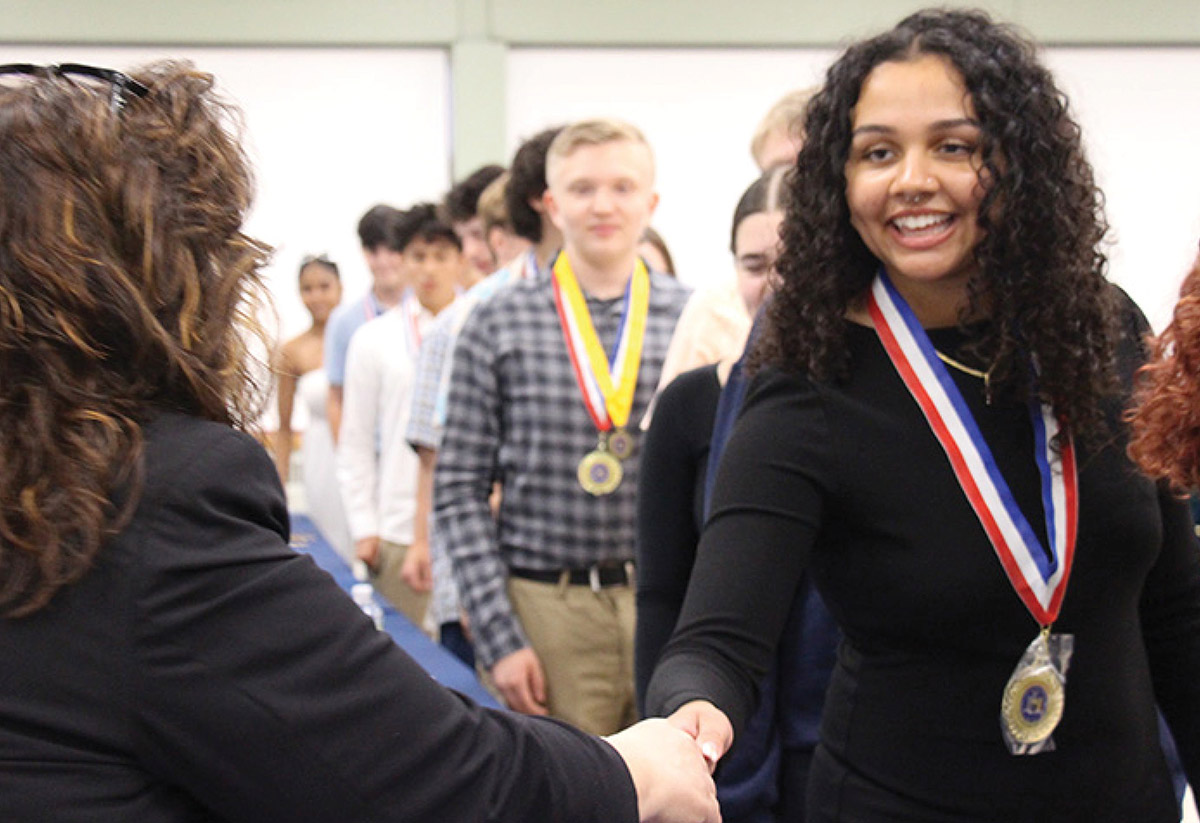 A smiling young woman with curly dark hair shakes hands at an award ceremony while wearing a medal on a red, white, and blue ribbon. Other students wearing medals stand in a line behind her.