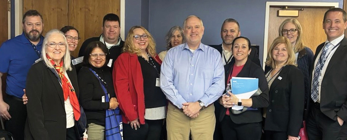 A group of professionally dressed men and women smiling and posing together indoors against a blue wall, with a man in a light blue button-down shirt standing at the center.