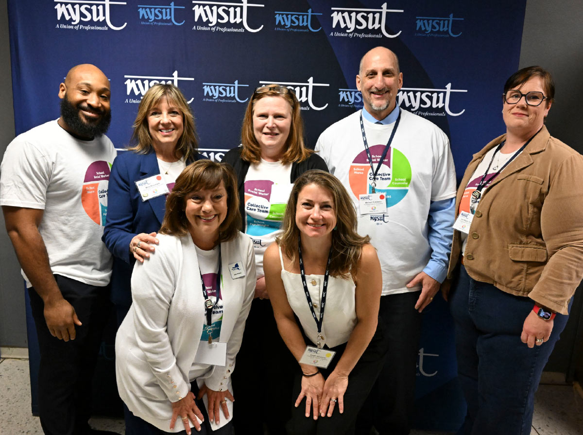 A group of six smiling professionals poses together in front of a blue backdrop with the NYSUT union logo repeating.