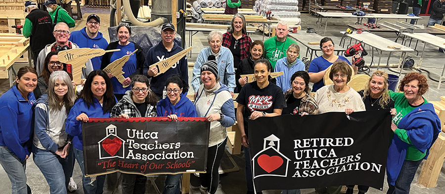 A large group of volunteers holding Utica Teachers Association banners and wooden cutouts in a workshop.