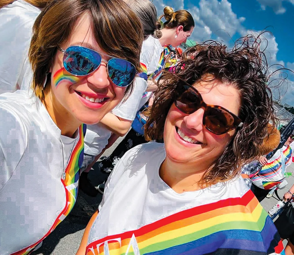 Two smiling women in sunglasses wear rainbow face paint and sashes at an outdoor Pride parade celebration.