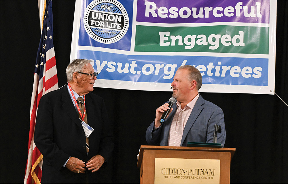 NYSUT Vice President Ron Gross speaks into a microphone at a podium, introducing award-recipient Paul Cole.