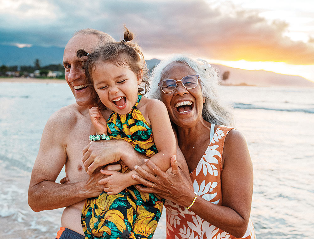 Grandparents laughing and holding a young girl on a beach during sunset.