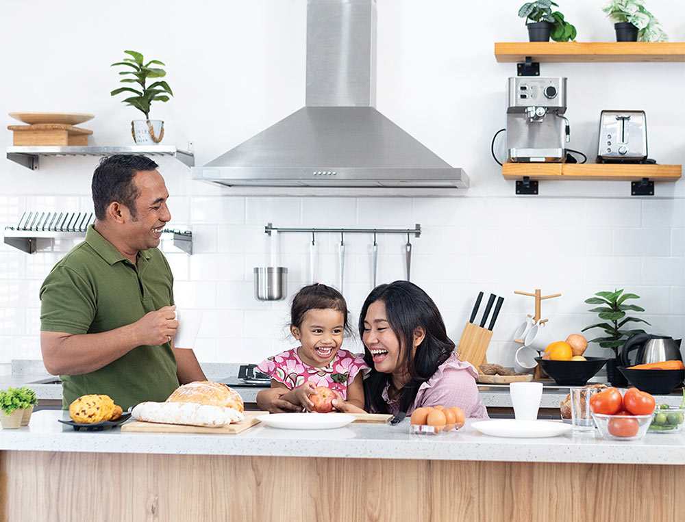 A man and woman laugh with a young girl as all three stand in a kitchen preparing a meal together.