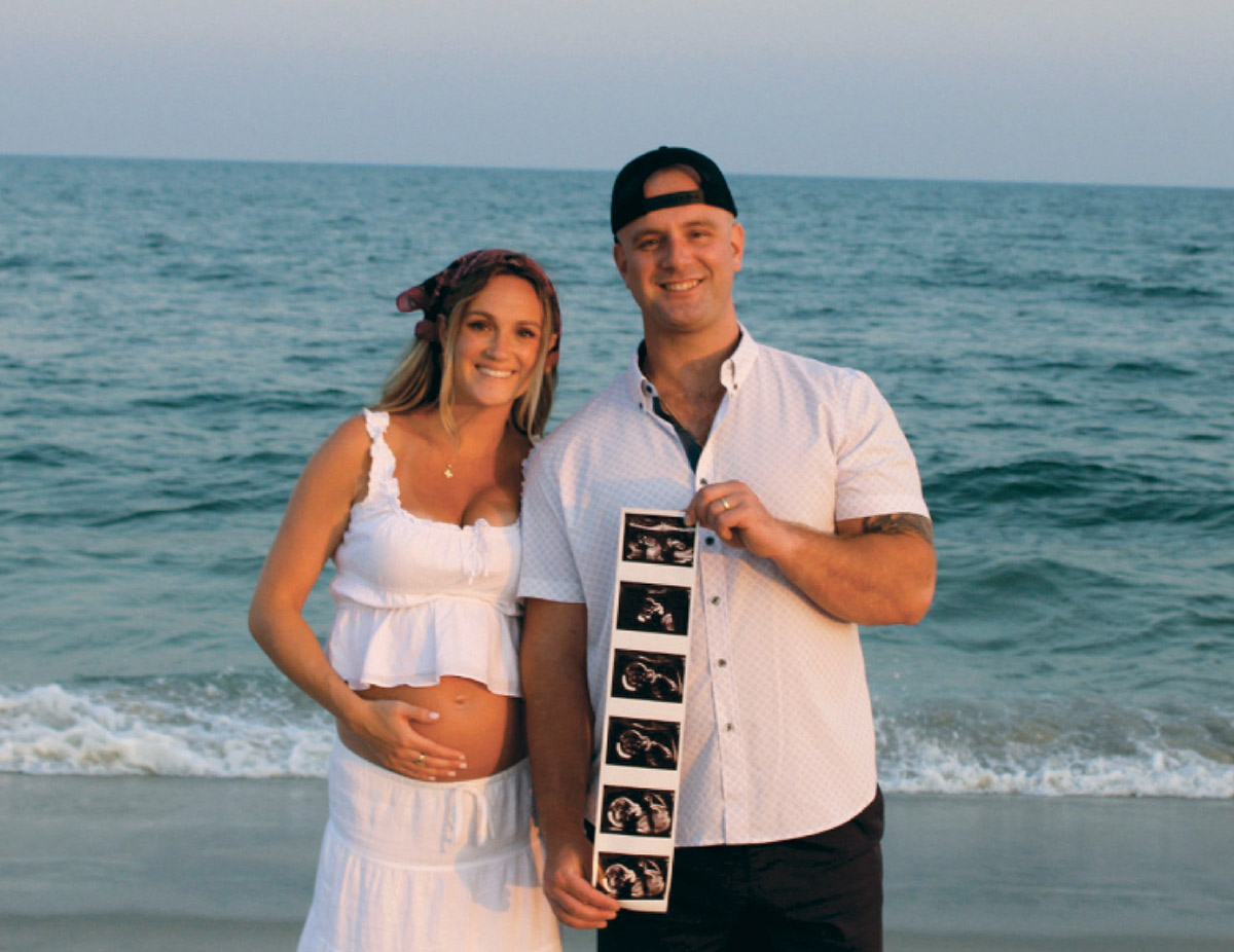 Smiling expectant couple standing on a beach holding a strip of ultrasound photos.