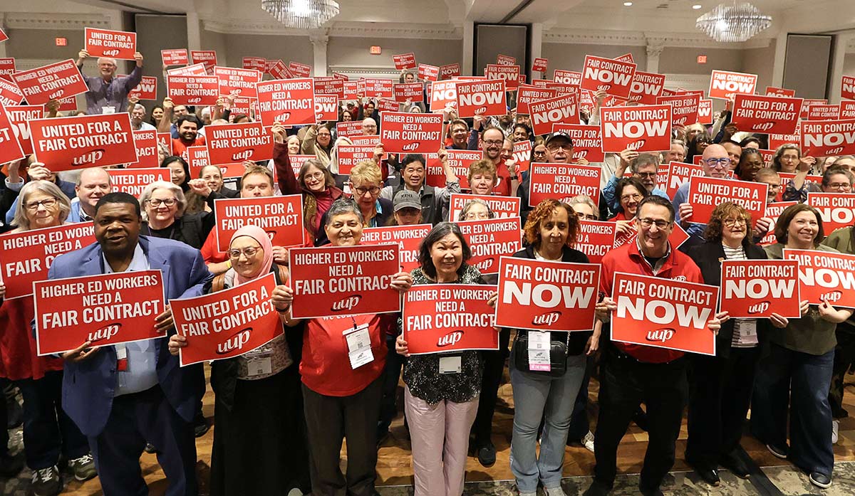 Crowd of union members holding red "Fair Contract Now" signs at an indoor rally.