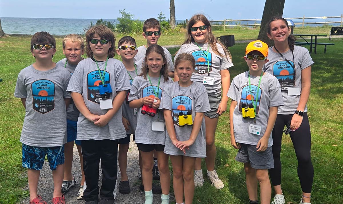A group of children and an instructor wearing matching "Guardian of Lake Ontario" t-shirts and binoculars pose outdoors near the water.