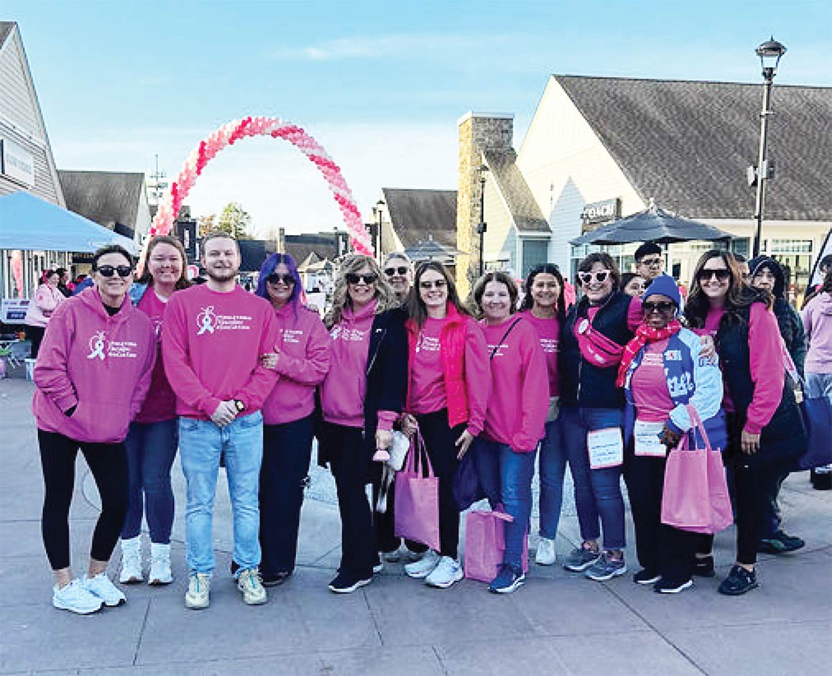 A group of participants wearing matching pink shirts pose together outdoors in front of a pink balloon arch at a breast cancer awareness walk.
