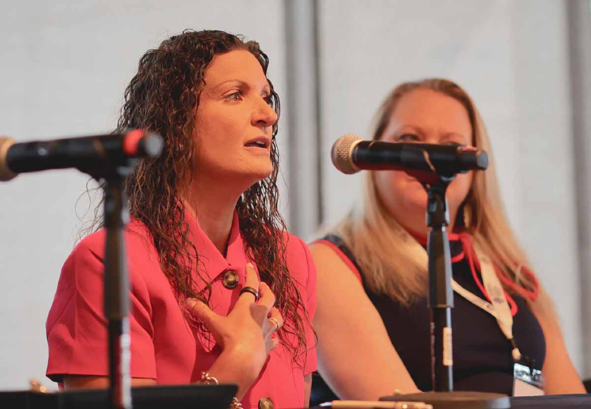 Woman with curly hair in a pink jacket speaking passionately into a microphone seated beside a colleague during a panel.