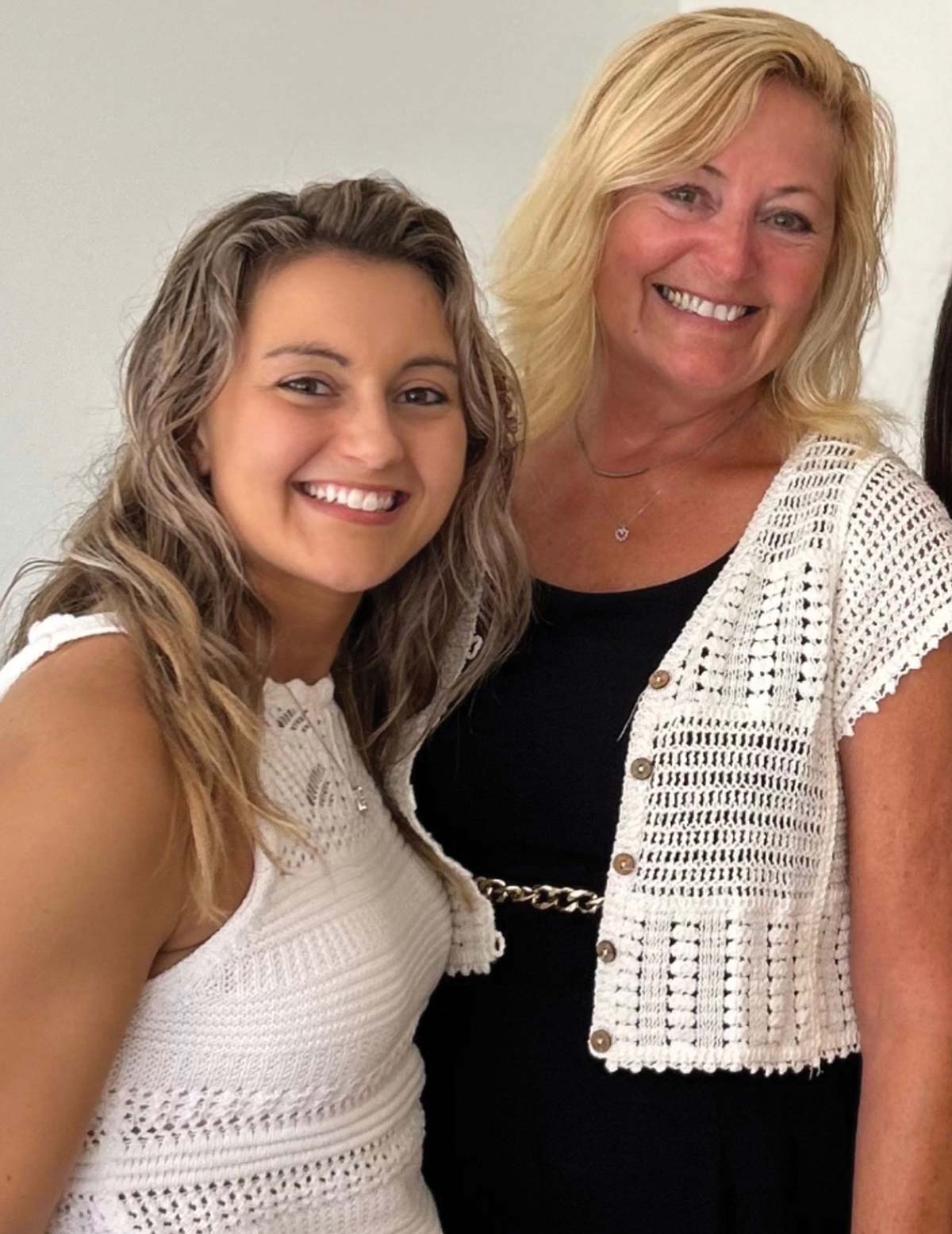 Two women smiling together for a photo, one wearing a white knit top and the other wearing a black top with a white crocheted cardigan.