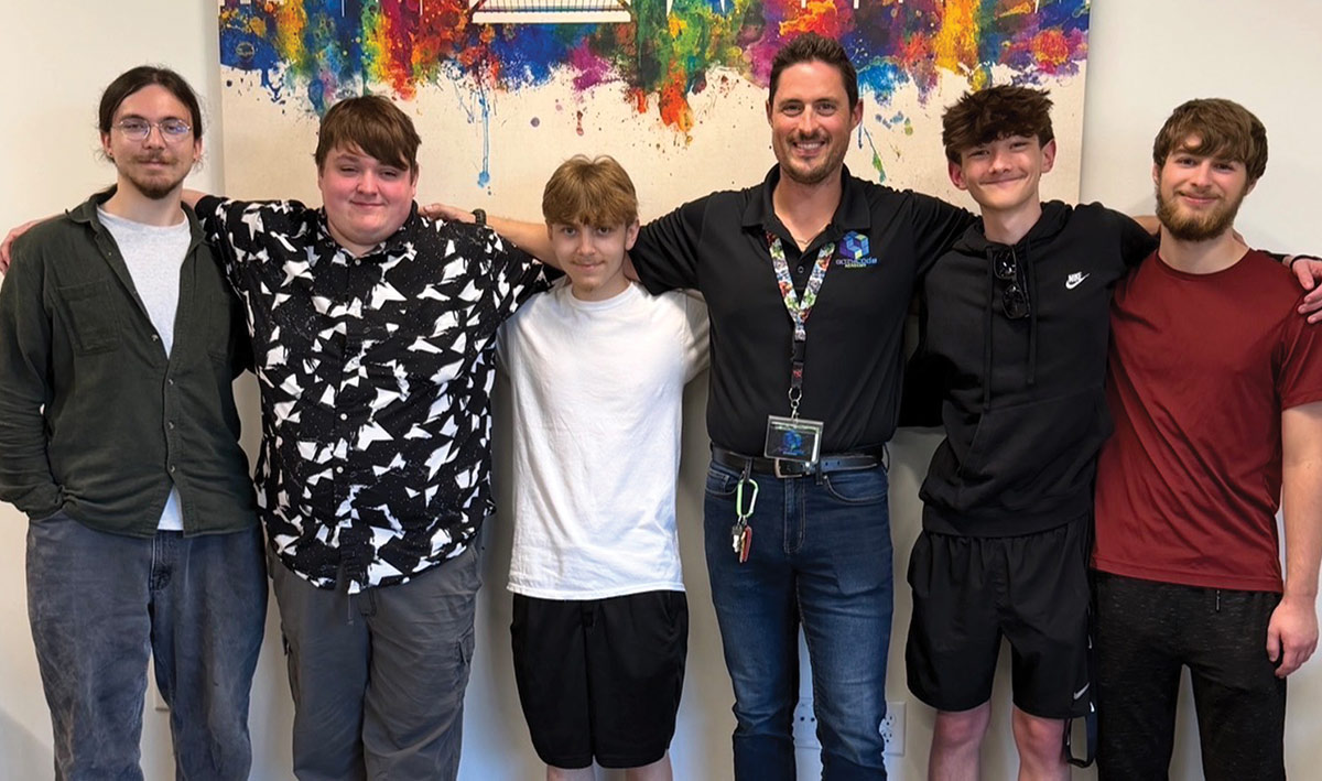An instructor wearing a lanyard posing with five young men standing in a line with their arms around each other in front of colorful wall art.