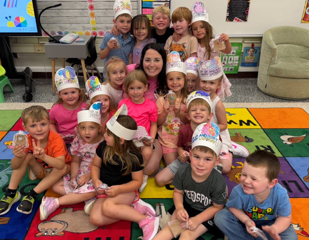 Teacher posing on a colorful classroom rug with a group of smiling young students wearing decorated paper crowns.