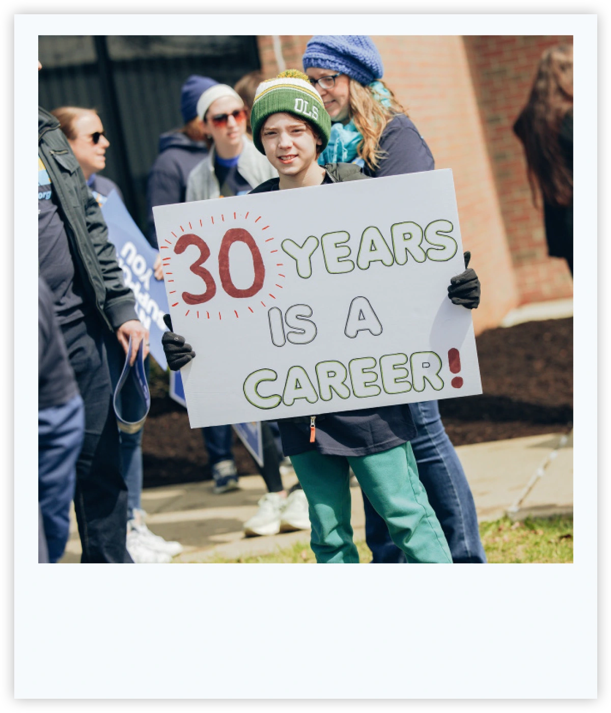 Young person in a green beanie holding a handmade protest sign reading "30 Years Is a Career!"