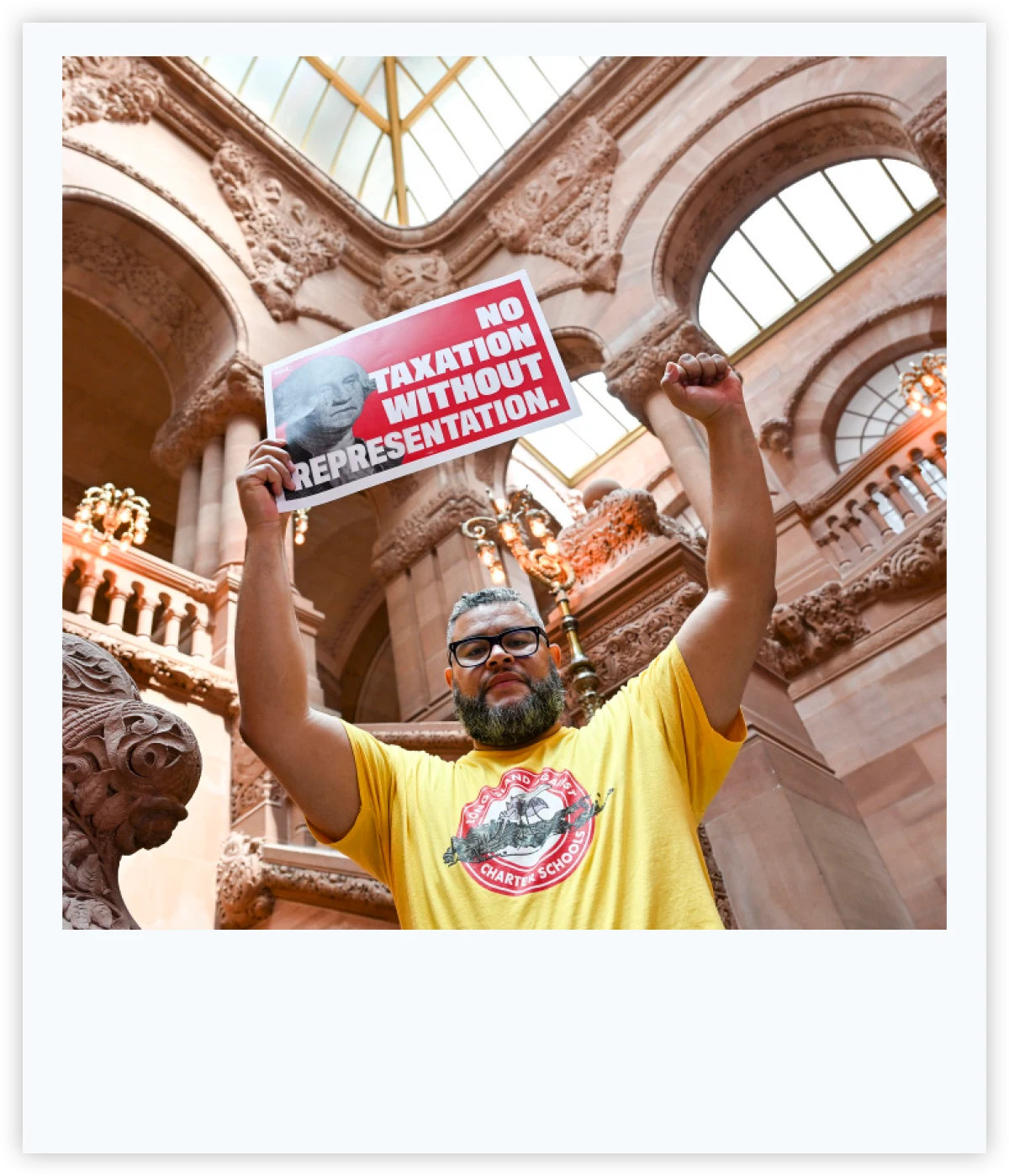 Man raising a fist inside a government building while holding a red sign reading "No Taxation Without Representation."