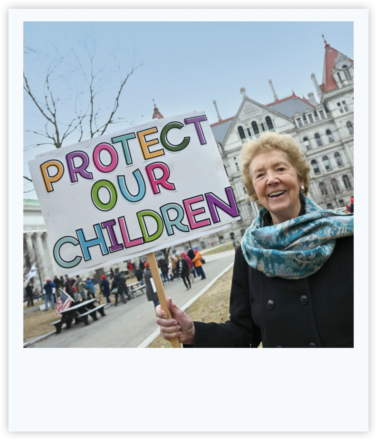 Smiling woman outdoors holding a sign with colorful lettering that reads "Protect Our Children."