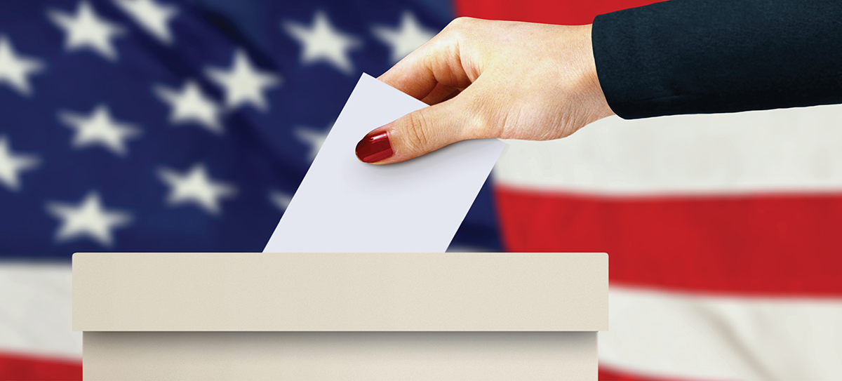 Hand inserting a paper ballot into a ballot box in front of an American flag background.